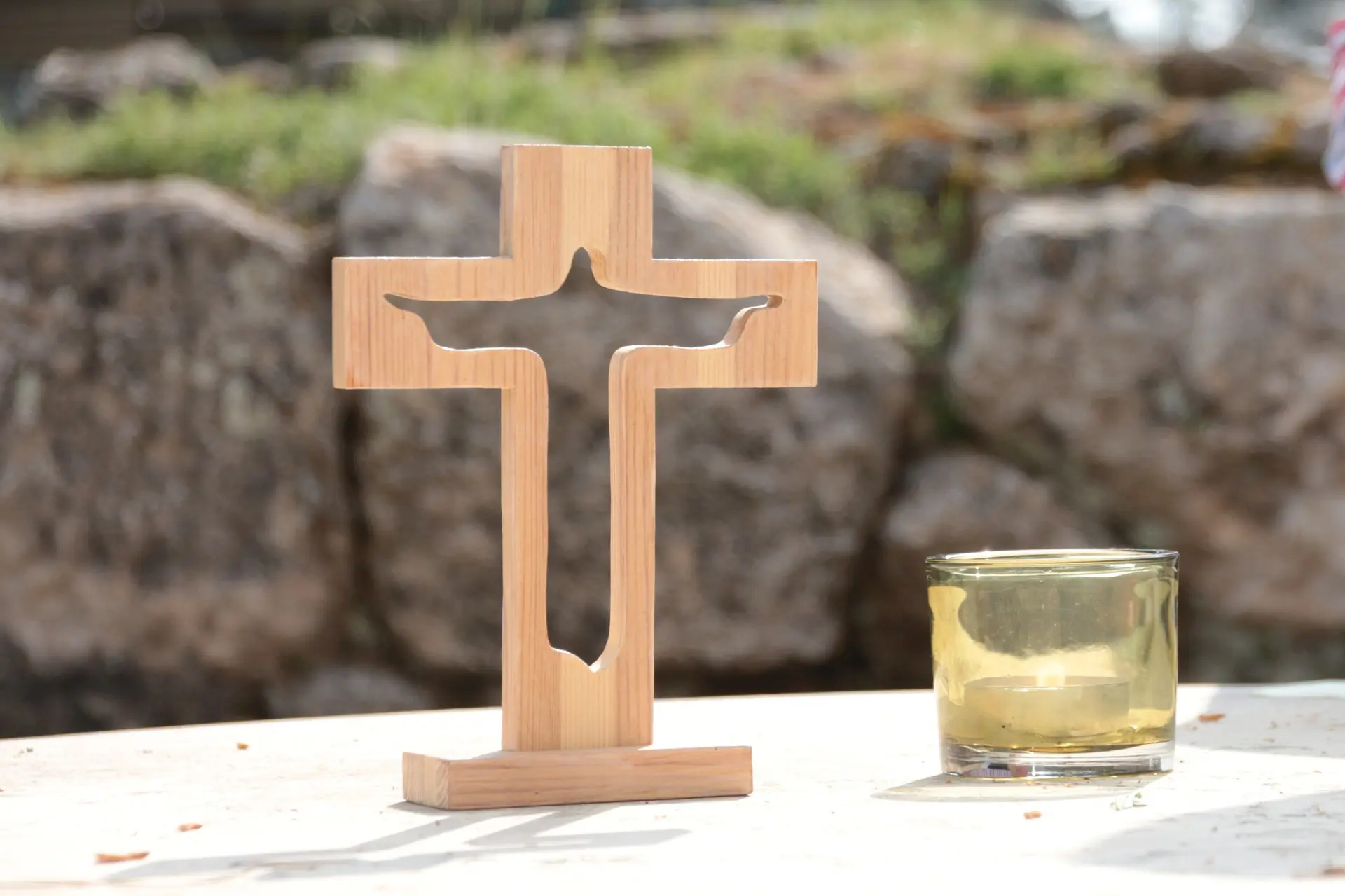 Ein Holzkreuz, aus dem eine Jesus-Figur ausgeschnitten ist, steht auf eine glatten Oberfläche. Daneben ein brennendes Teelicht in einem gelben Glas. Der Hintergrund besteht aus eine undeutlich sichtbaren Mauer.